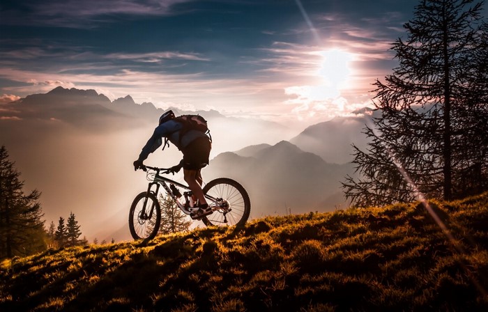 A cyclist rides downhill through a scenic mountain trail with dramatic sunset lighting.