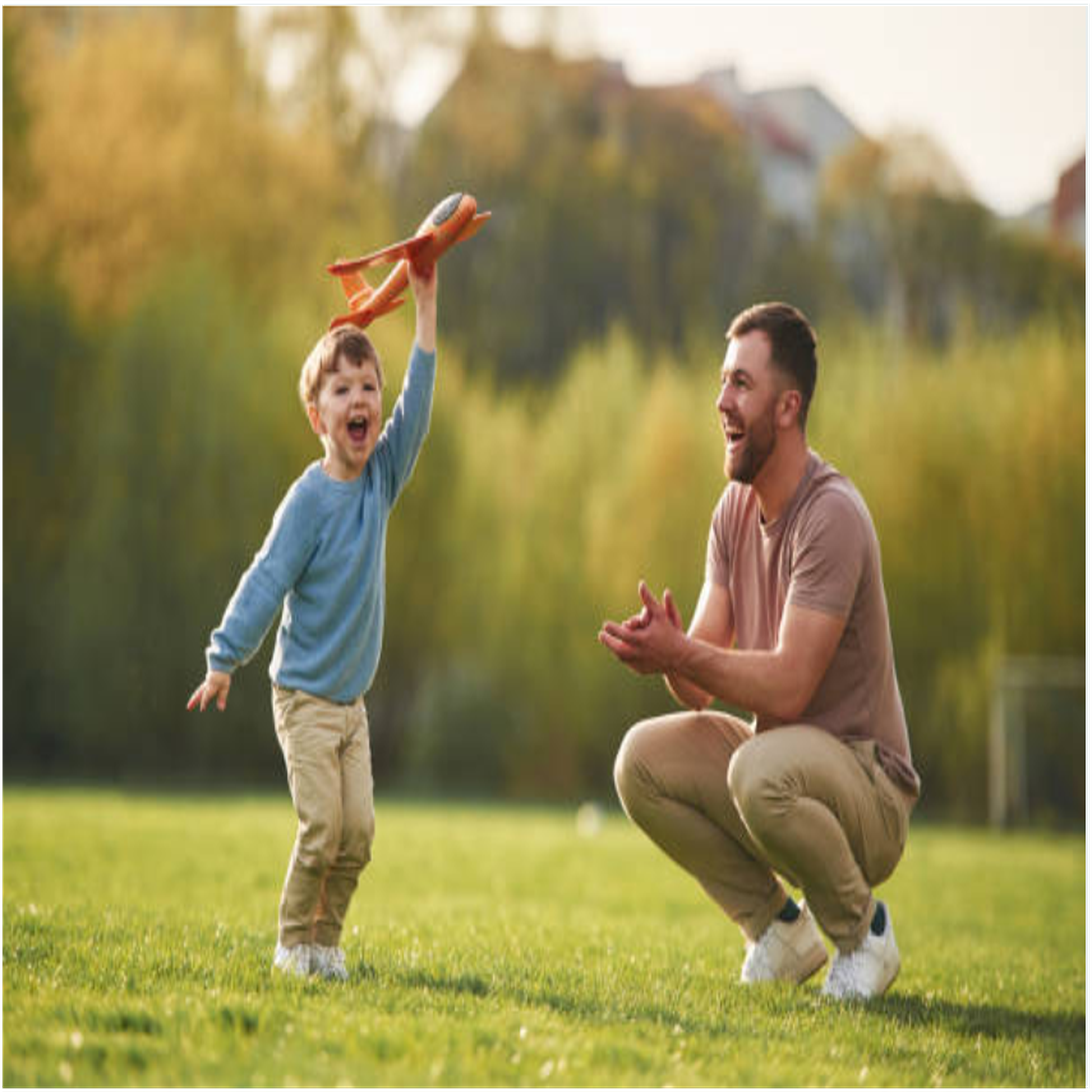 A joyful father watches his son holding a toy airplane in a green park with autumn trees behind them. A joyful father watches his son holding a toy airplane in a green park with autumn trees behind them.
