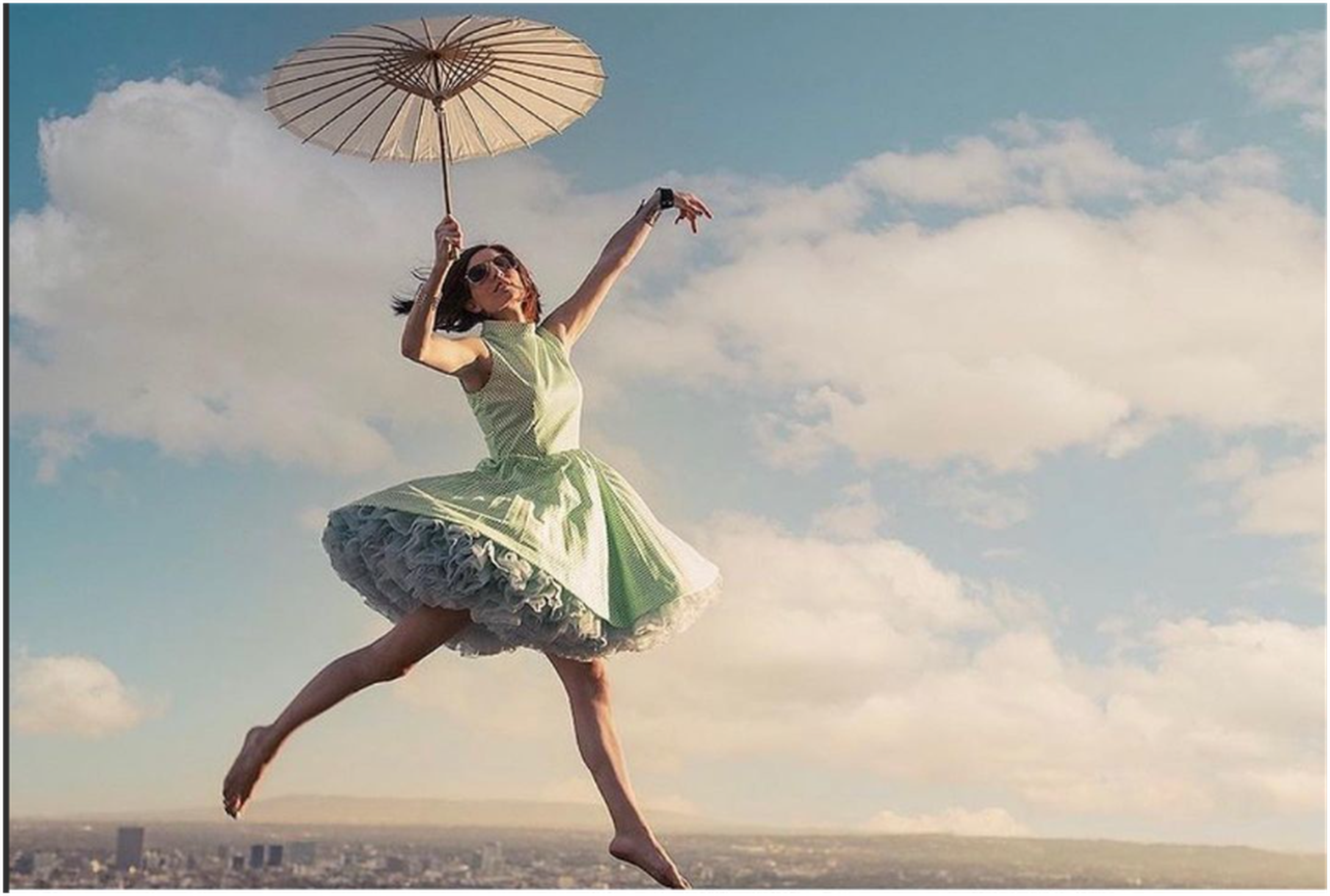 A woman in a dress and sunglasses appears to float mid-air holding a parasol against a cloudy sky. A woman in a dress and sunglasses appears to float mid-air holding a parasol against a cloudy sky.